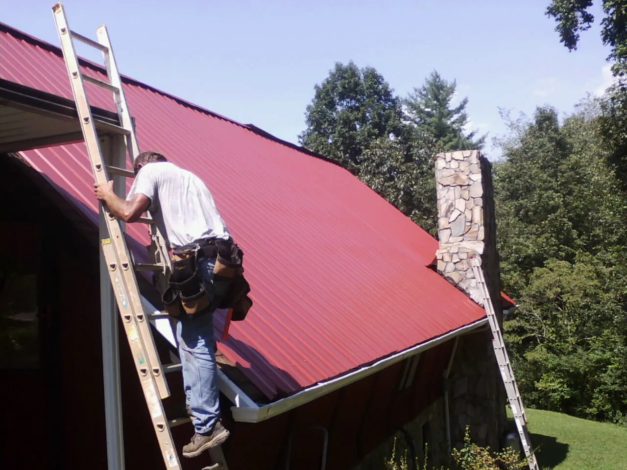 Roofer on a ladder beside a red metal roof and stone chimney — Soffit & Fascia Repair by Roofing Brothers in Closter